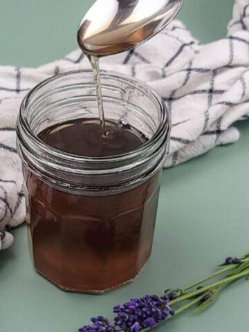 A spoon drizzles honey into a jar on a green surface. A white cloth with a black grid pattern is placed beside the jar, and a small bunch of lavender is positioned in the foreground.