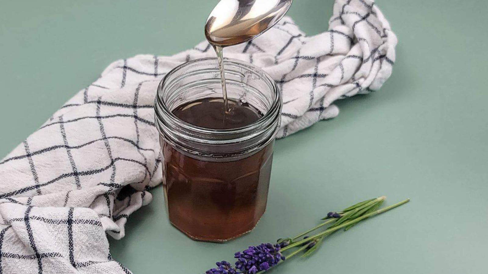 A spoon drizzles honey into a jar on a green surface. A white cloth with a black grid pattern is placed beside the jar, and a small bunch of lavender is positioned in the foreground.