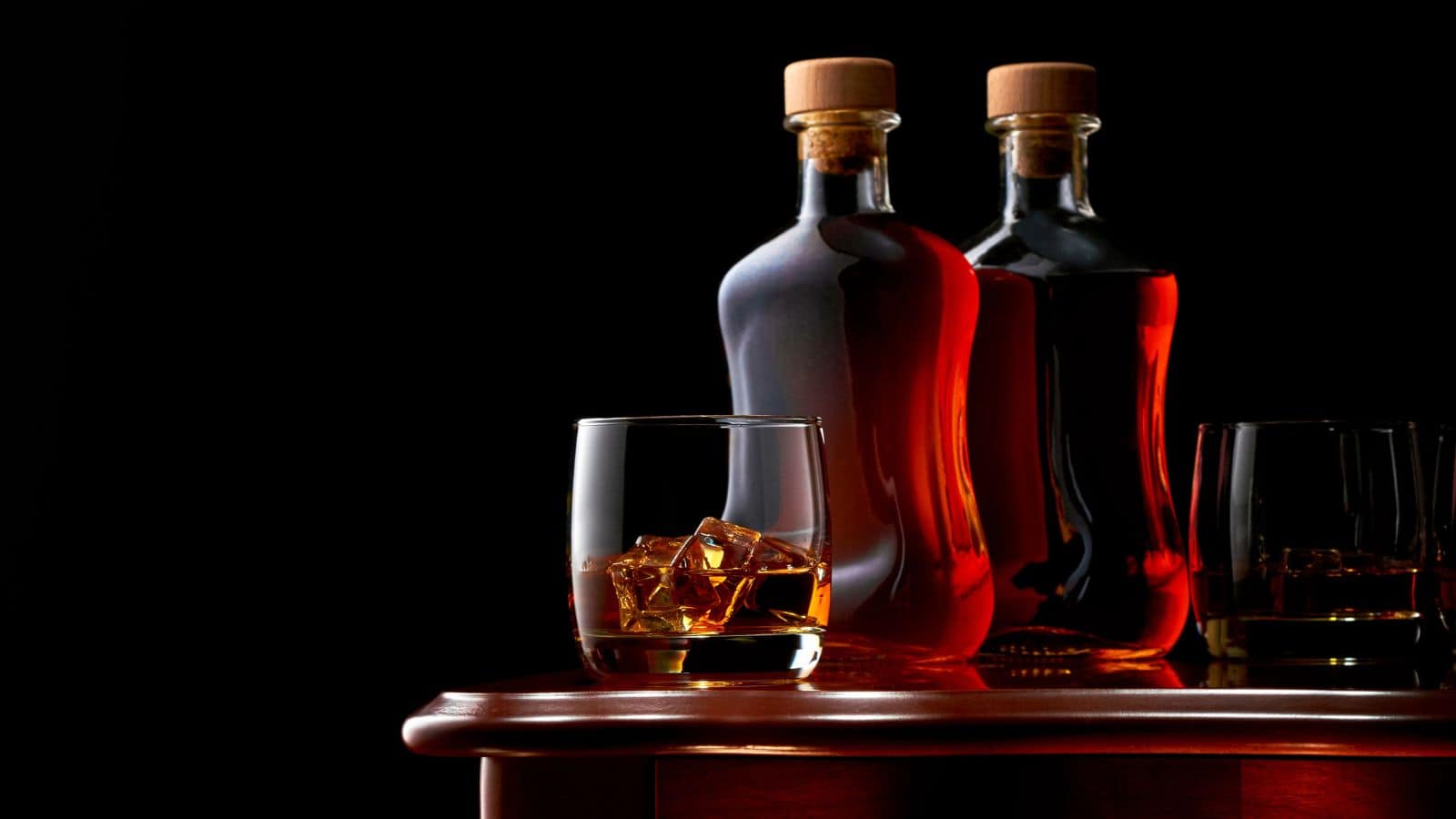 Two glass bottles filled with dark liquor stand on a wooden surface against a black background. In the foreground, a glass with ice cubes contains a serving of the liquor. Another similar glass is partially visible on the right.