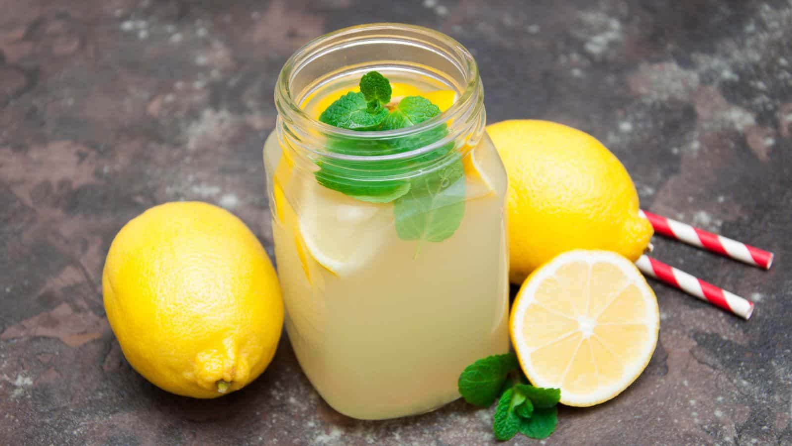 A glass jar filled with lemonade, garnished with lemon slices and mint leaves, is placed on a textured surface. Two whole lemons, half a lemon, and red striped straws are beside the jar.