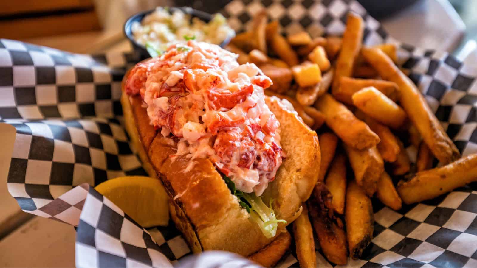 A lobster roll filled with lobster meat is placed on a checkered paper-lined basket. It is accompanied by golden-brown French fries and a lemon wedge. A small container of coleslaw is visible in the background.
