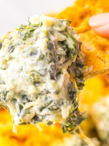 A hand holding a cracker dipped in a creamy spinach and cheese mixture, with a baked dish of the same mixture in the background.