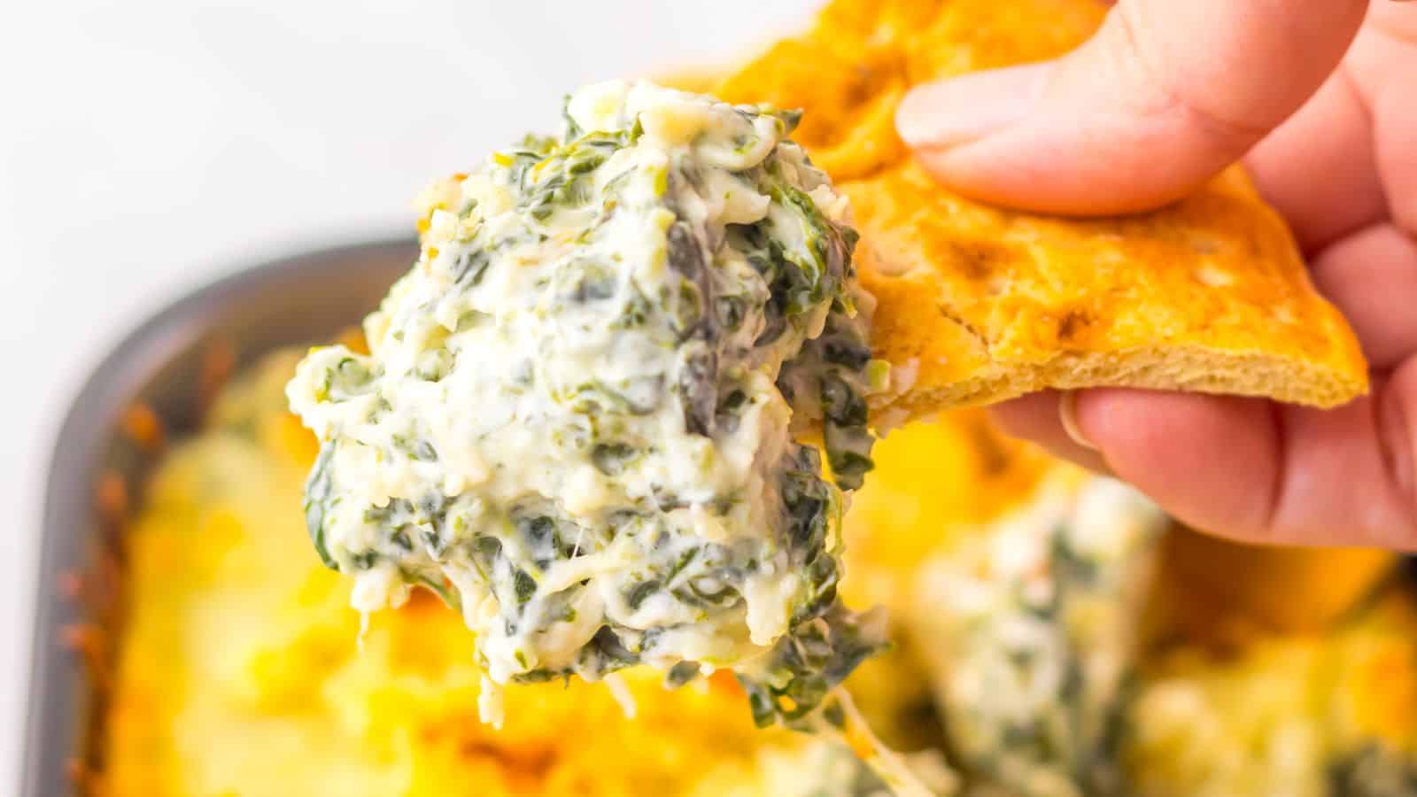 A hand holding a cracker dipped in a creamy spinach and cheese mixture, with a baked dish of the same mixture in the background.