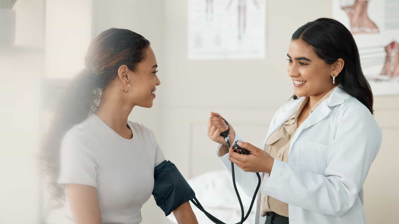 A healthcare professional takes a blood pressure reading of a seated patient. The patient wears a black cuff on her arm, and both are engaged in conversation in a medical office setting. Diagrams are visible on the wall in the background.
