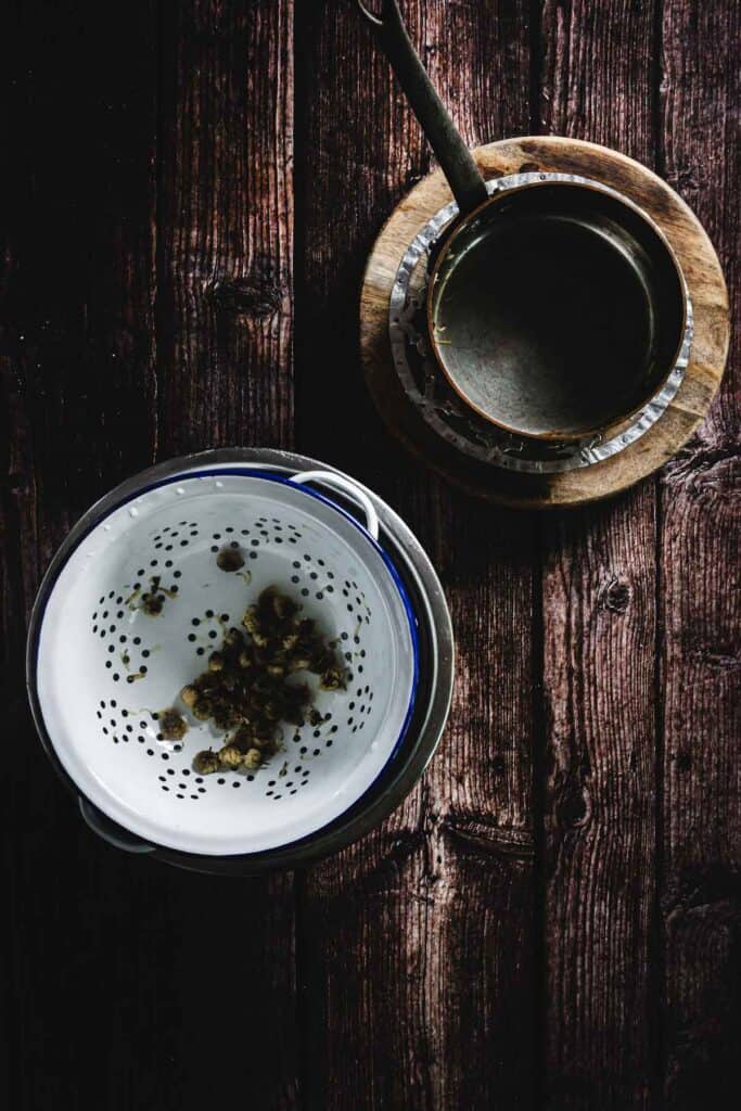A white colander with holes contains small brown objects, possibly spices or seeds. Next to it is a small saucepan on a round wooden board. Both are placed on a dark wooden surface.