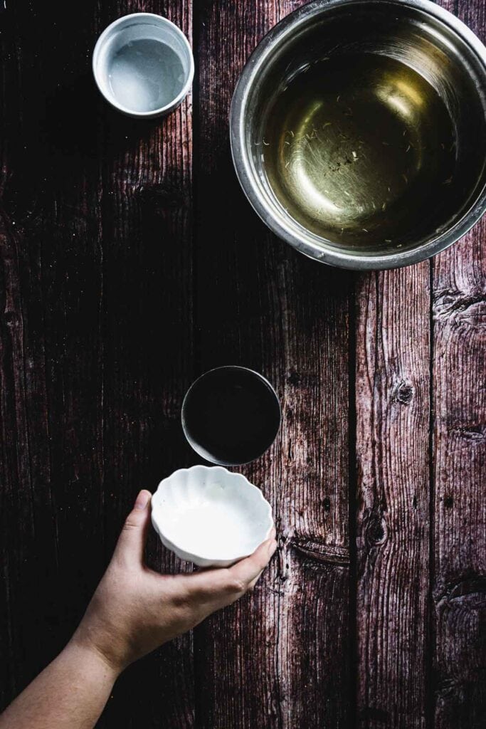 A hand holds a small white bowl over a wooden table. Nearby, there are two other bowls and a large metal mixing bowl containing a yellow liquid.