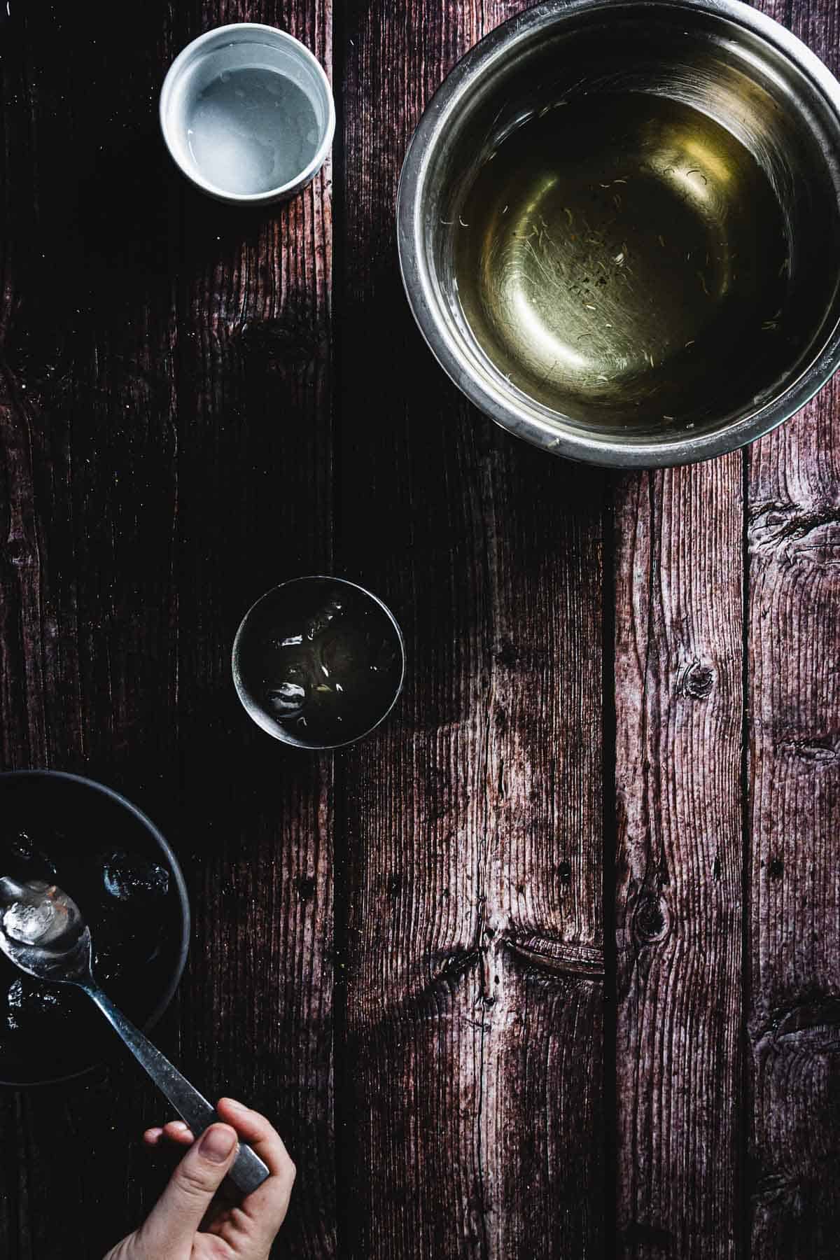 A wooden table featuring a metal bowl with liquid, a white cup containing a small amount of liquid, a dark bowl with ice cubes and a spoon, and a hand holding the spoon. The table surface is dark and textured with visible wood grain.