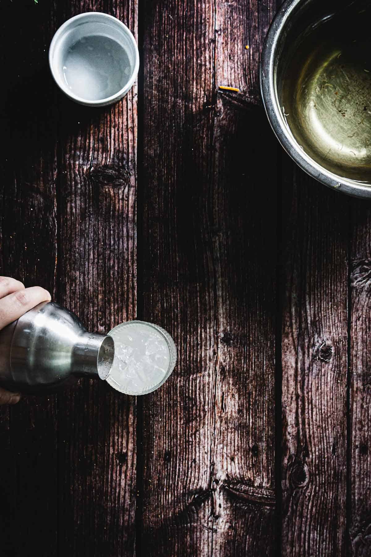 A hand pours a clear liquid from a shaker into a glass on a rustic wooden surface. Two bowls, one empty and one with liquid, are nearby.