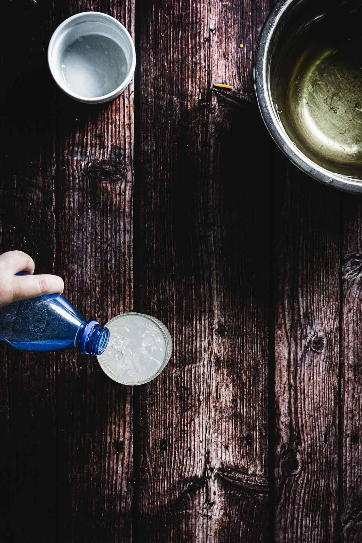 A hand pours clear liquid from a blue bottle into a glass on a rustic wooden table. Nearby are an empty white cup and a silver bowl. The scene captures a moment of drink preparation or kitchen activity.