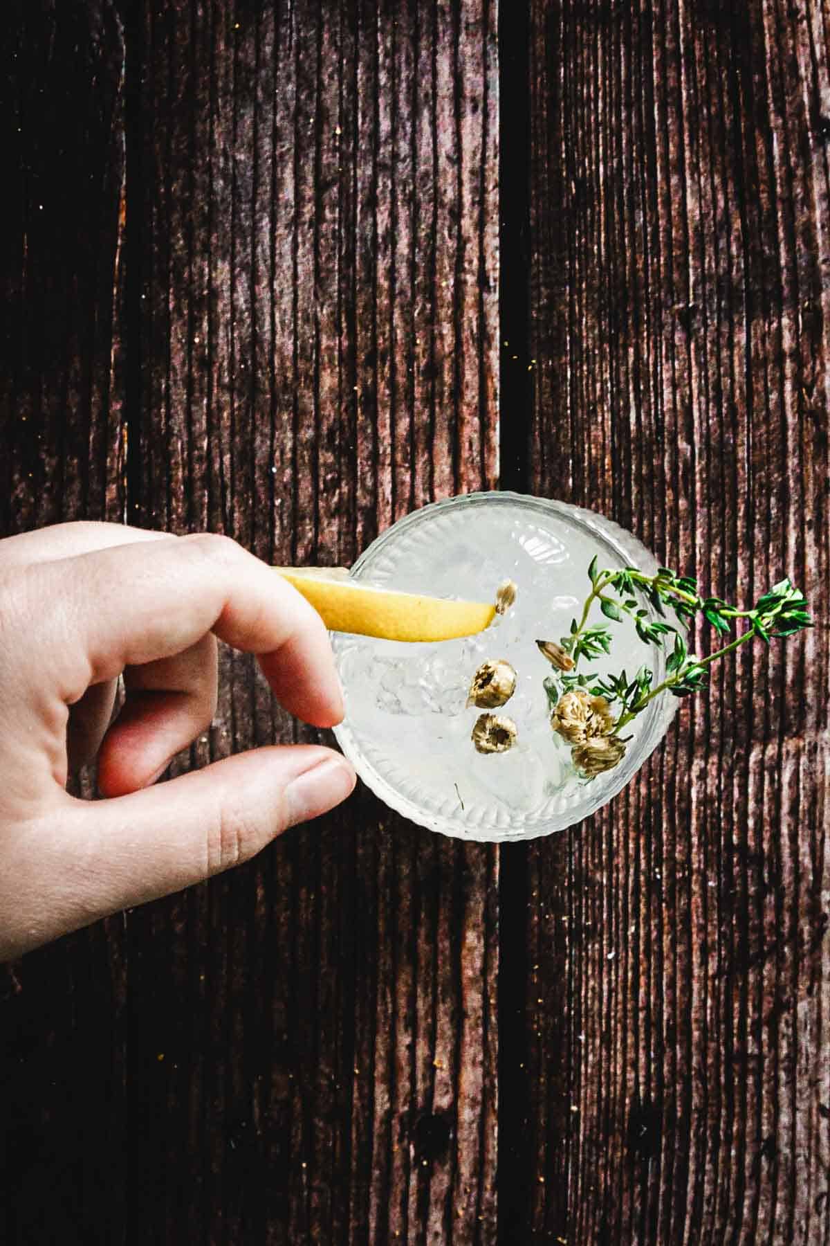A hand holds a clear cocktail glass containing a light beverage garnished with a lemon twist, green herbs, and a few small round objects. The drink rests on a dark, textured wooden surface.