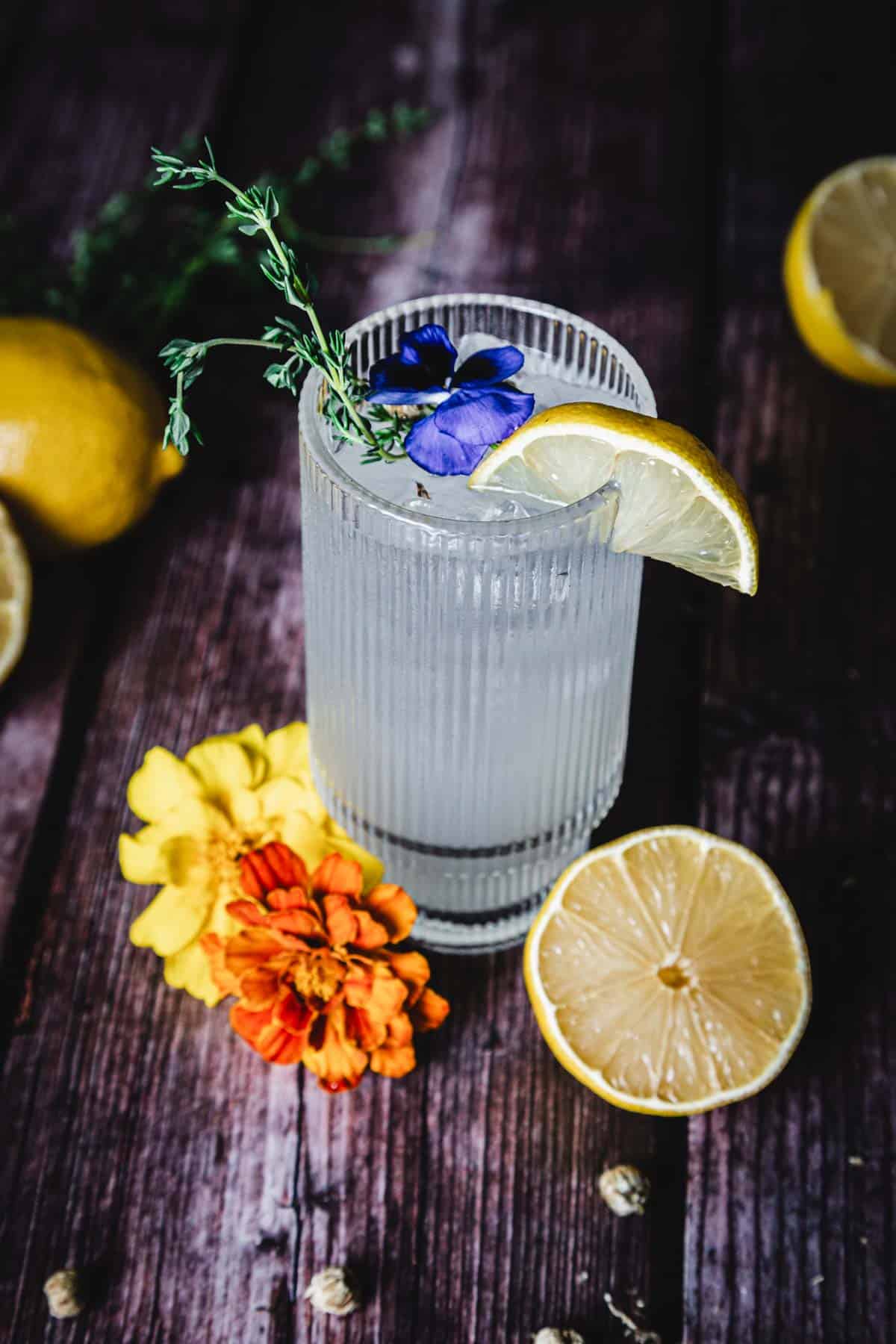 A glass of clear liquid with ice is garnished with a lemon slice, a purple flower, and a sprig of herbs. It's placed on a wooden surface, surrounded by half a lemon and two vibrant flowers, one orange and one yellow.
