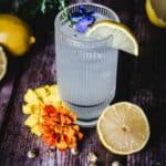 A textured glass filled with a clear beverage, garnished with a lemon slice, a sprig of herbs, and a blue flower. Surrounding the glass are lemons, a sliced lemon, two orange and yellow flowers, and scattered seeds on a wooden surface.