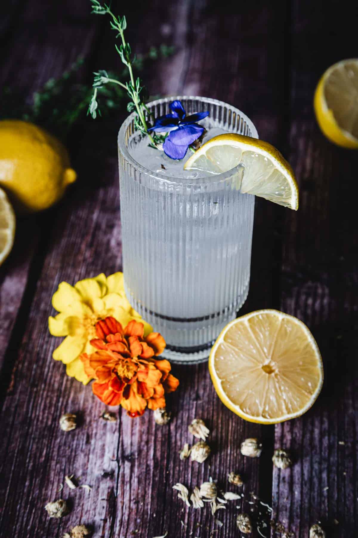 A textured glass filled with a clear beverage, garnished with a lemon slice, a sprig of herbs, and a blue flower. Surrounding the glass are lemons, a sliced lemon, two orange and yellow flowers, and scattered seeds on a wooden surface.