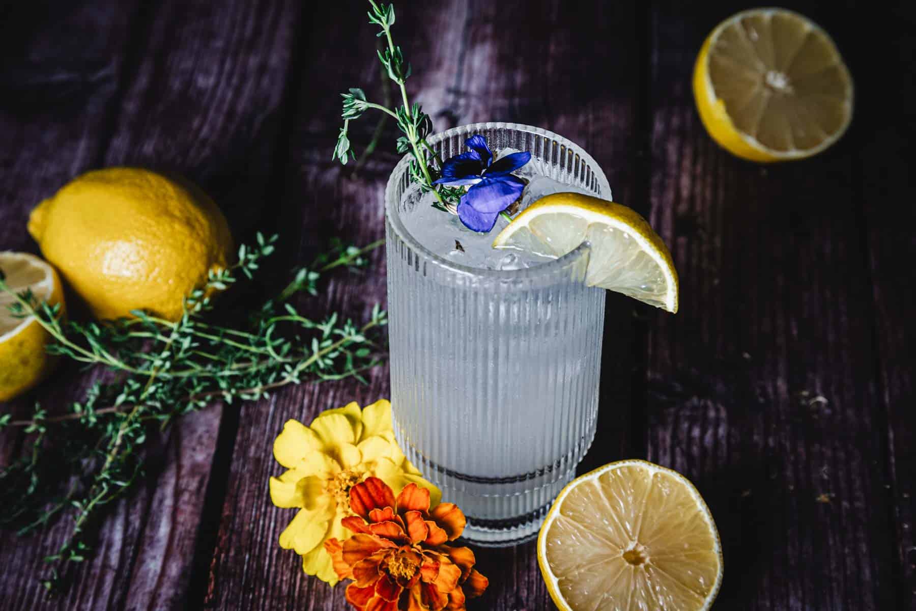 A glass of clear cocktail garnished with a lemon slice, blue flower, and herbs sits on a dark wooden table. Surrounding the glass are whole and halved lemons, sprigs of greenery, and yellow and orange flowers.