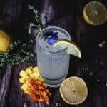 A tall, ribbed glass filled with ice and a clear beverage, garnished with a lemon slice, blue flower, and green sprig. Surrounding the drink are yellow and orange flowers, a sprig of herbs, a whole lemon, and a halved lemon on a dark wooden surface.