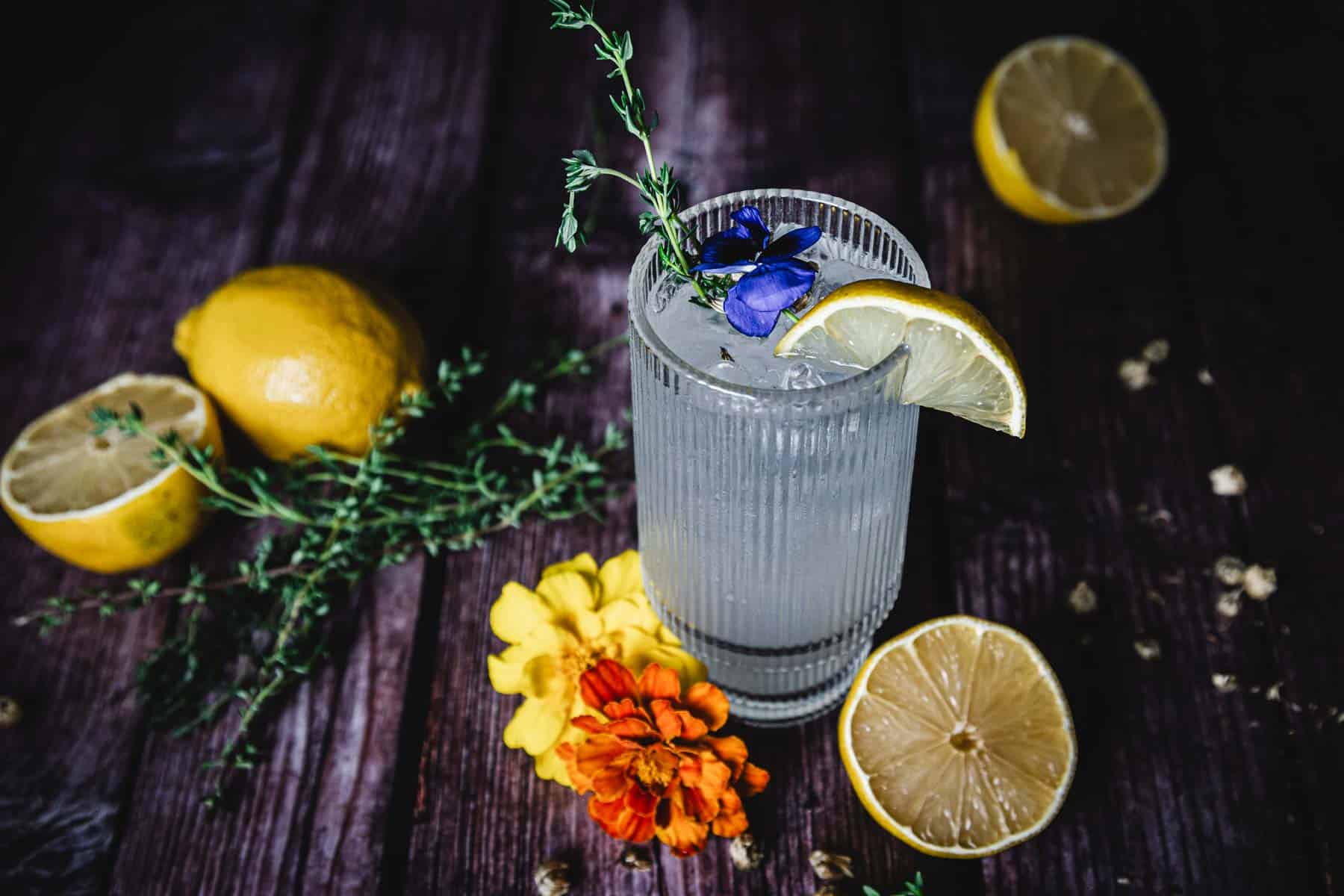 A glass of lemonade garnished with a lemon slice and sprigs of greenery, placed on a wooden surface. Surrounding the glass are whole and sliced lemons, small flowers, and scattered pieces of greenery.