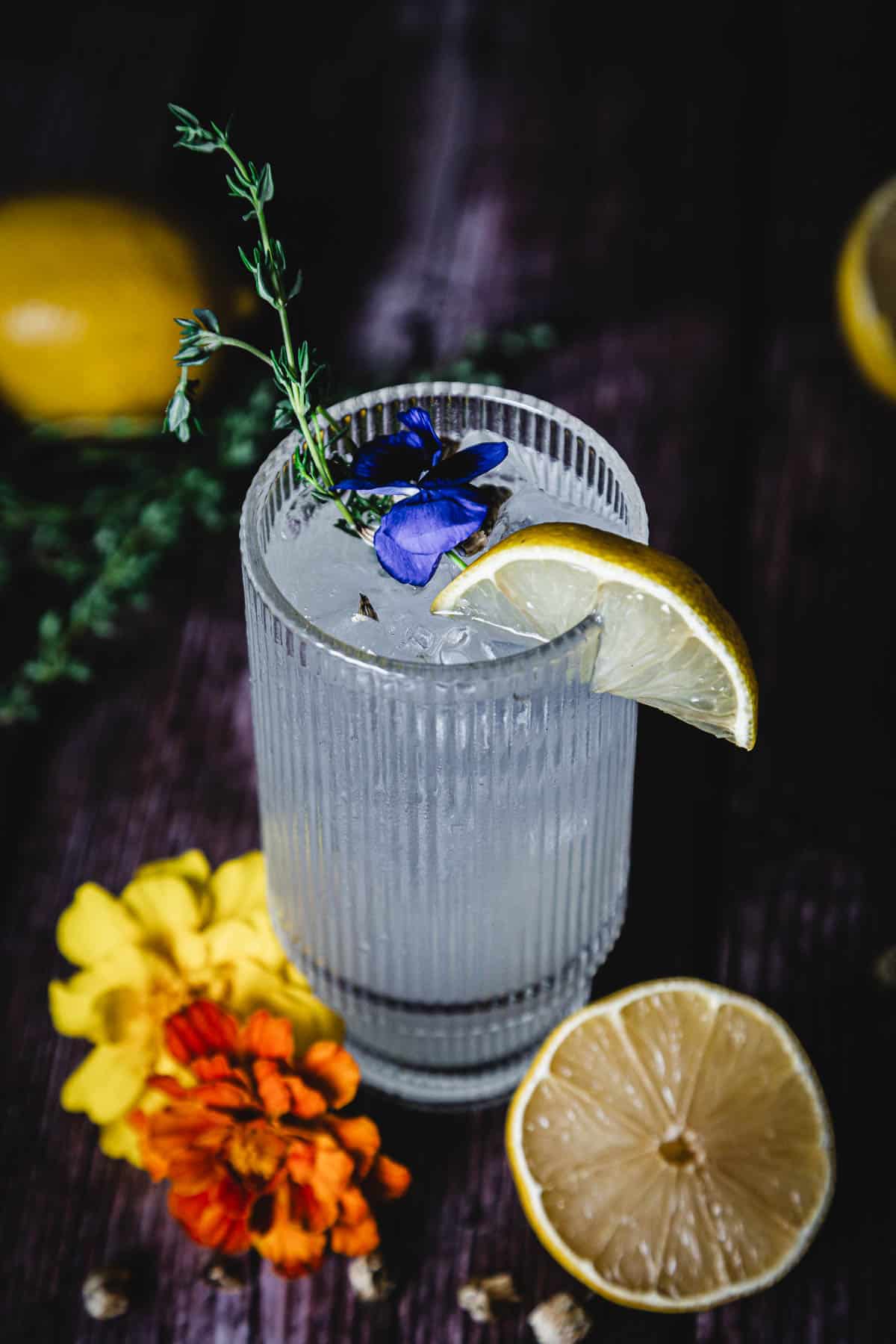 A glass of a clear cocktail garnished with a lemon slice, a sprig of thyme, and a purple flower. The glass is on a dark wooden surface, surrounded by yellow and orange flowers, and lemon halves.