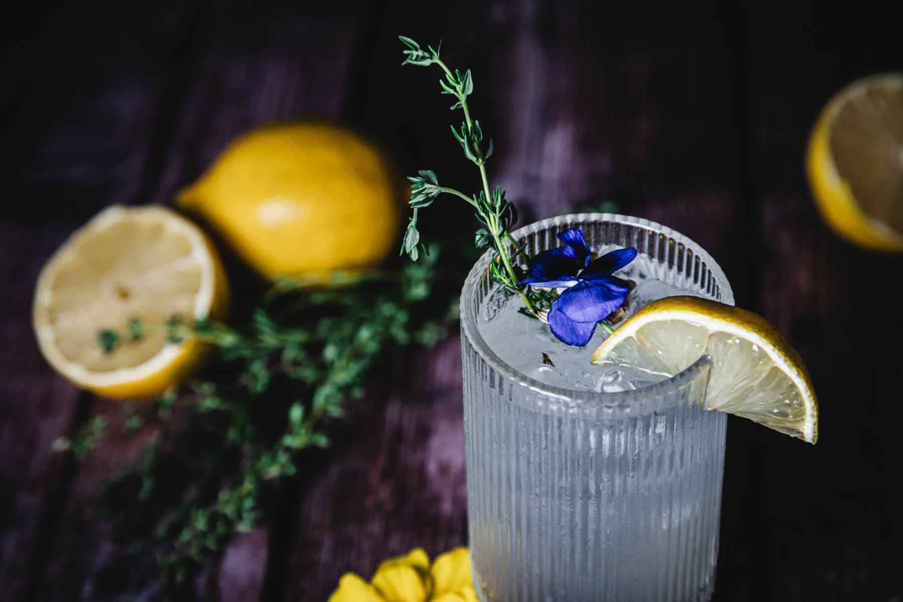 A textured glass of iced beverage is garnished with a lemon slice, a sprig of herbs, and a purple flower. In the background, there are whole and halved lemons on a dark wooden surface.