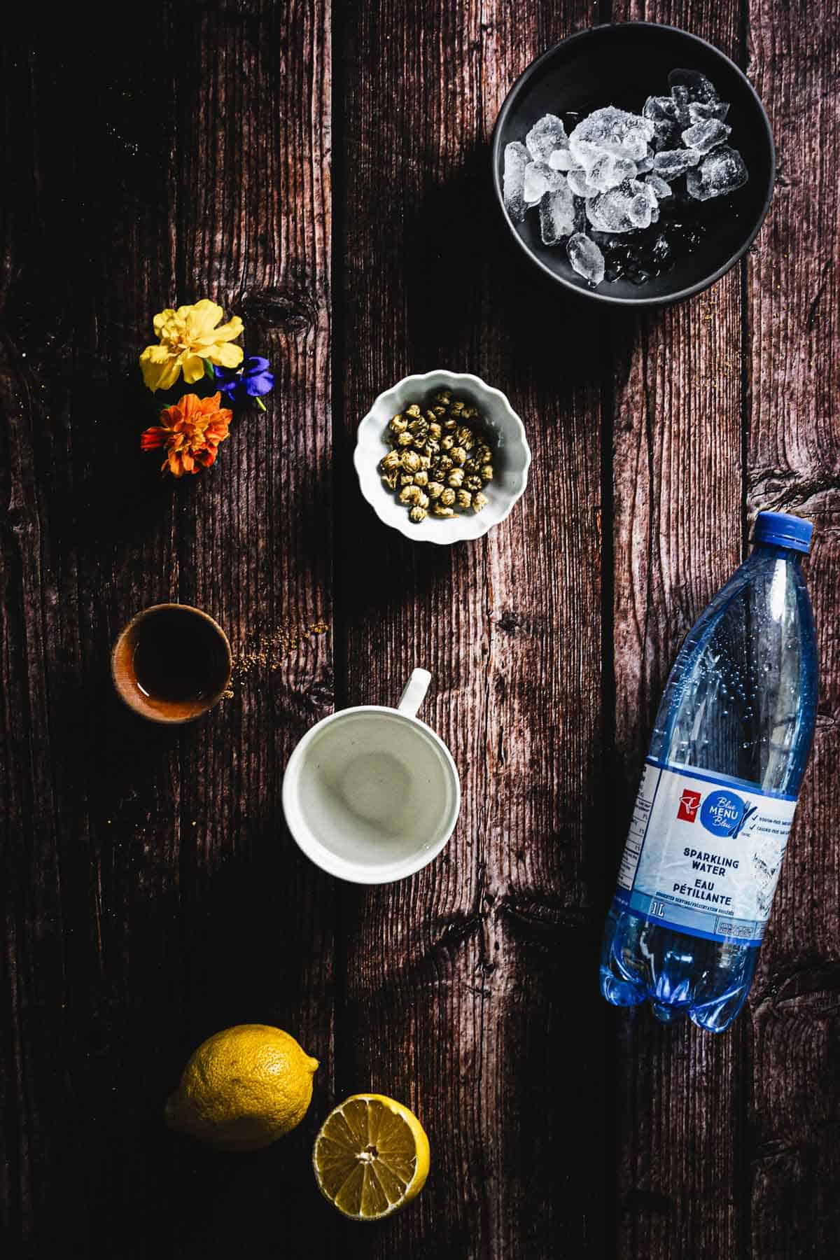 A wooden table displays a bottle of sparkling water, a mug, a small bowl of capers, ice cubes in a black bowl, a halved lemon, and a few colorful flowers. A small cup is also present, possibly containing sauce or spices.