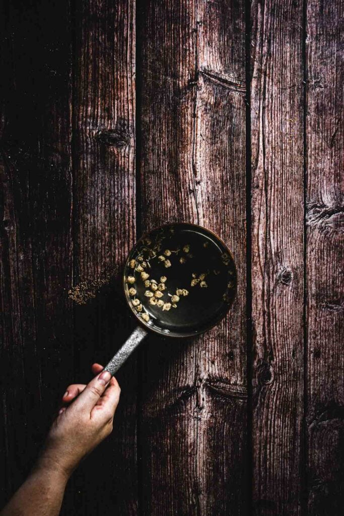 A hand holds a saucepan with a metal handle over a wooden surface. The saucepan contains some grains or seeds in a liquid, viewed from above. The wooden surface is dark and rustic.