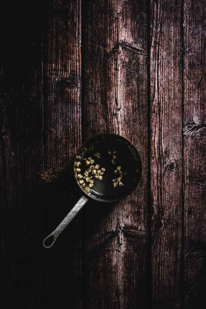 A pot with a metal handle contains water and dried chamomile, sitting on a dark, rustic wooden surface. The lighting creates a contrast between the pot and the textured backdrop, reminiscent of an artisanal mocktail's visual appeal.
