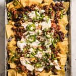 A baking tray filled with nachos topped with melted cheese, ground meat, kidney beans, sliced jalapeños, and a dollop of sour cream is placed on a marble surface.