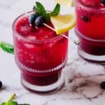 Two glasses of a deep red beverage garnished with lemon slices, mint leaves, and blueberries. The drinks are placed on a marble surface with additional mint leaves and blueberries scattered around.