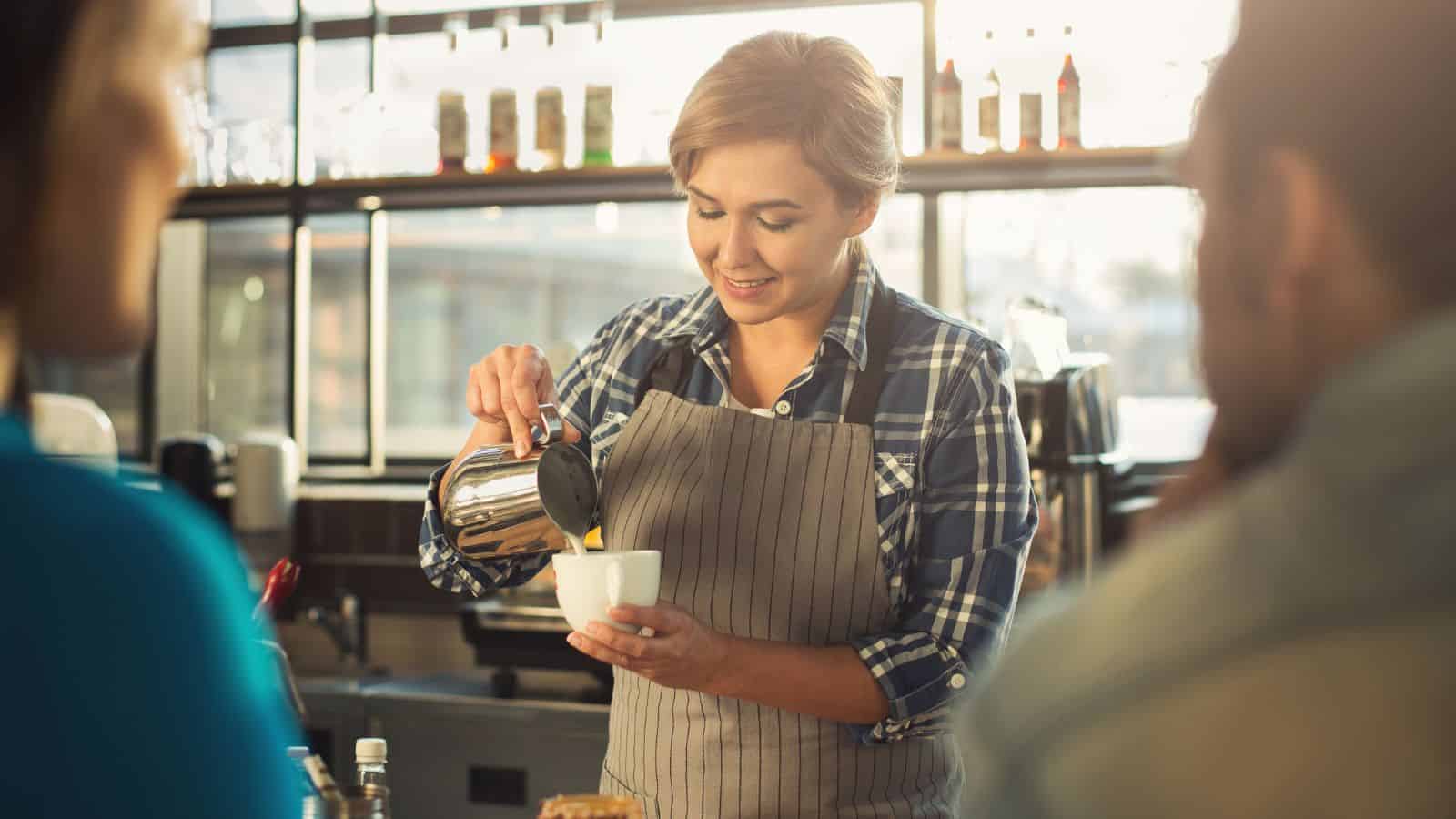 A barista in a plaid shirt and striped apron pours coffee from a pot into a cup. The setting is a well-lit café with bottles on a shelf in the background. Two people are seated at the counter in the foreground.