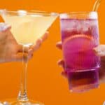 Two hands holding drinks in front of an orange background. The left hand holds a martini glass with a light yellow drink, while the right hand holds a tall glass with a pink drink and a straw.