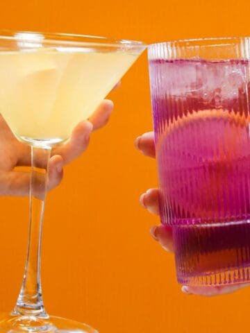Two hands holding drinks in front of an orange background. The left hand holds a martini glass with a light yellow drink, while the right hand holds a tall glass with a pink drink and a straw.