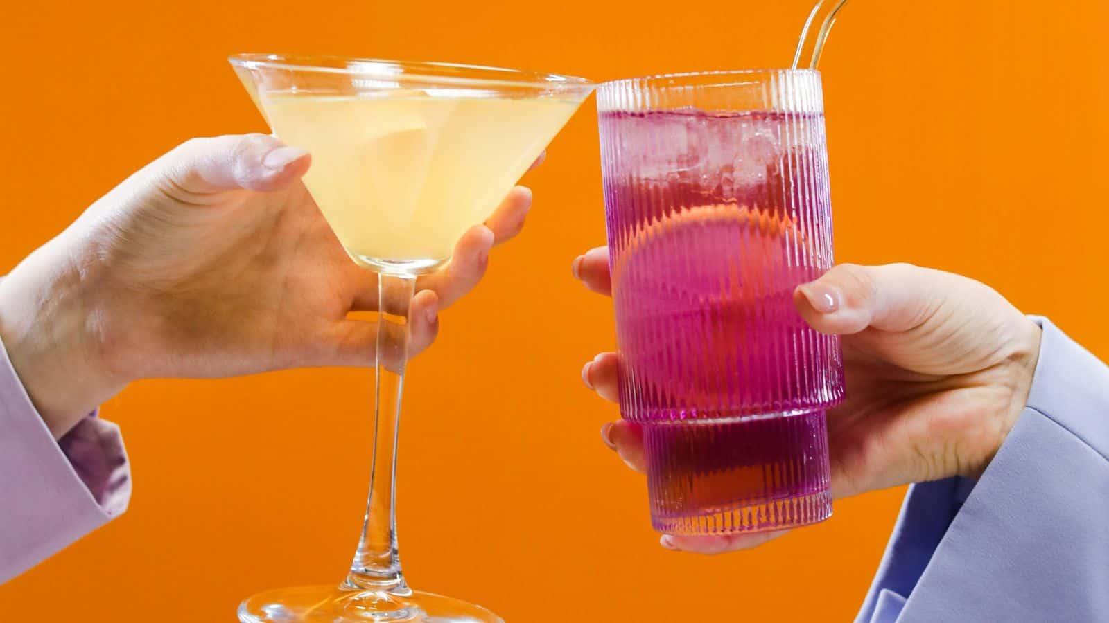 Two hands hold different drinks against an orange background. The left hand holds a yellow martini glass. The right hand holds a purple cocktail in a ribbed glass with a metal straw.