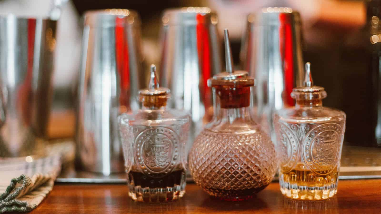 Three ornate glass bottles filled with different liquids are placed on a bar counter. Each bottle has a metal pourer. Shiny cocktail shakers are blurred in the background. The setting suggests a bar or cocktail-making station.