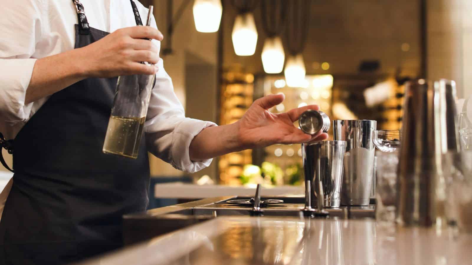 A person in an apron stands at a bar counter. They hold a bottle of liquid in one hand and a jigger in the other, pouring liquid into a shaker. The background shows blurred lights and shelves.