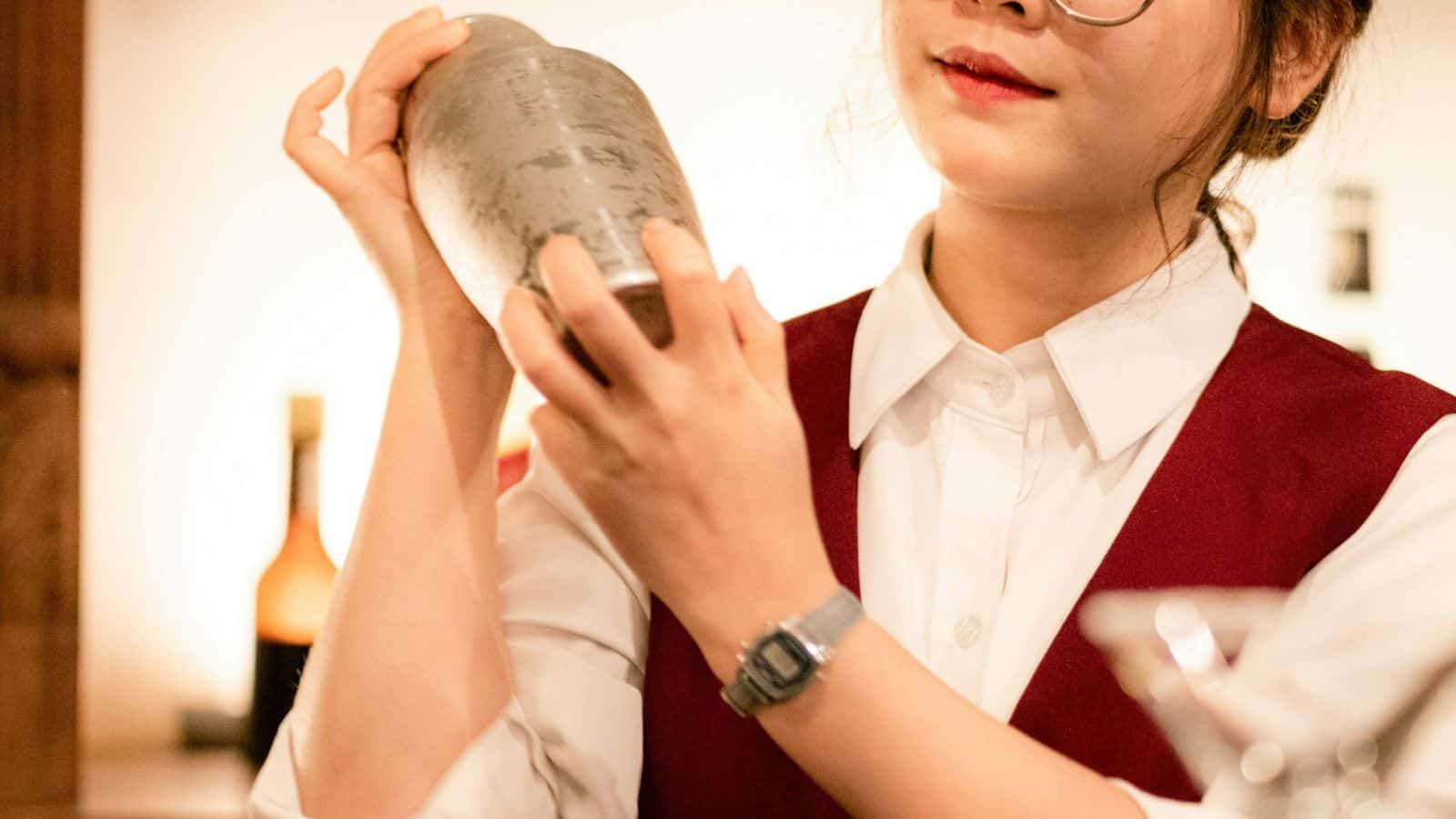 A person wearing glasses and a red vest over a white shirt is holding and shaking a metal cocktail shaker. Bottles and blurred background elements are visible.
