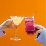 Two hands holding different drinks against an orange background: the left hand holds a martini glass with a light-colored beverage, and the right hand holds a tall glass with a purple drink and a metal straw.