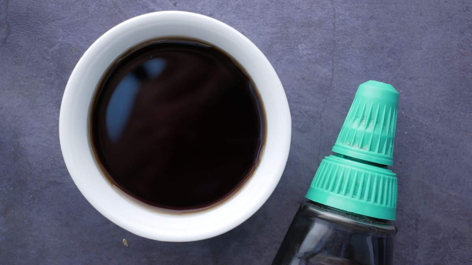 A white bowl filled with dark soy sauce is placed on a gray surface. Next to the bowl is a soy sauce bottle with a turquoise-green cap, positioned diagonally in the image.