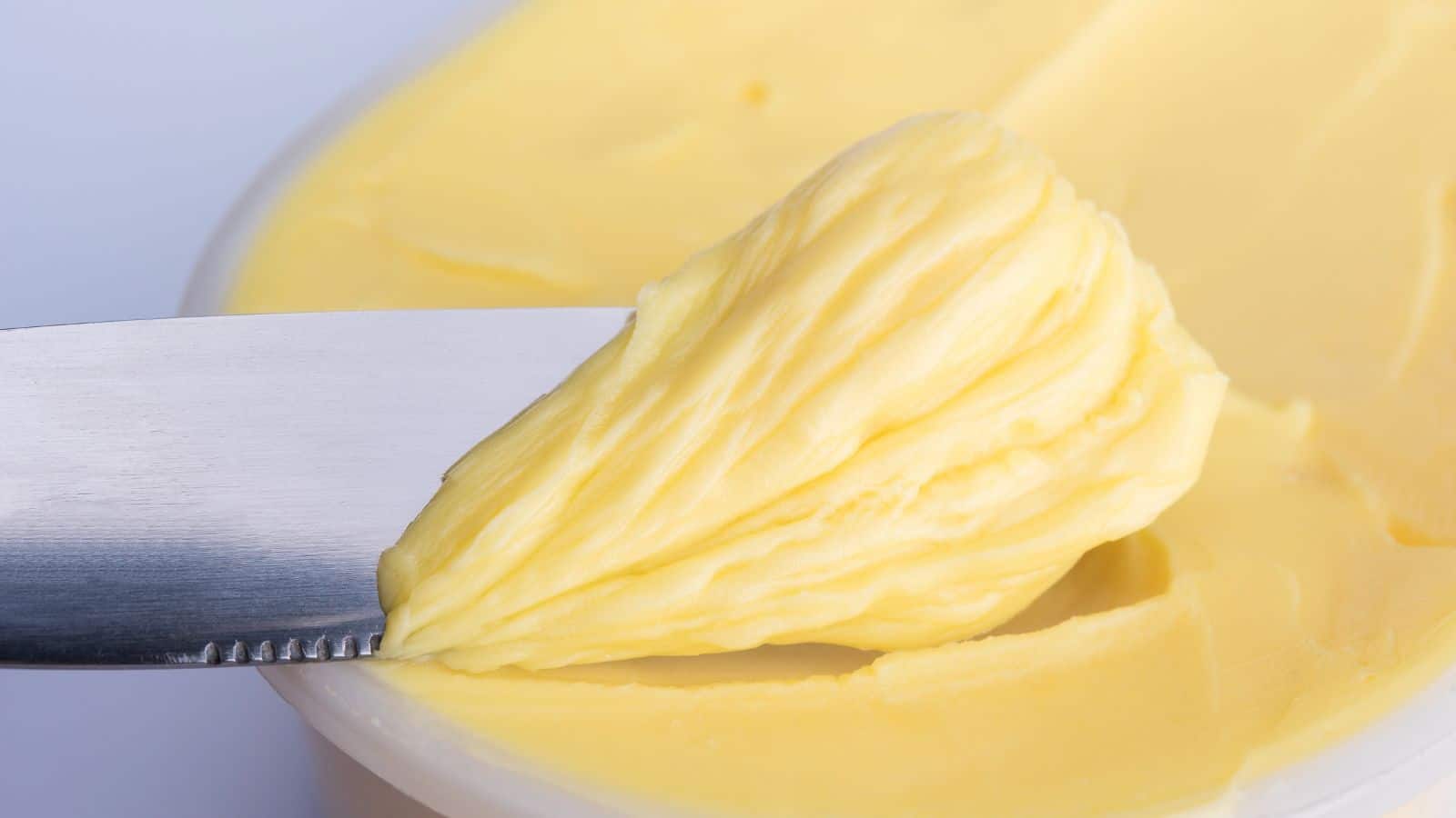 Close-up of a metal knife scooping a portion of smooth, pale yellow butter from a container. The butter has a soft, creamy texture, with ridges formed by the movement of the knife. The background is filled with more of the same butter.