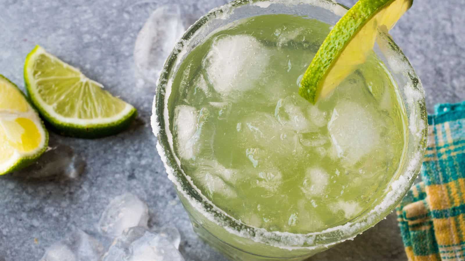A glass filled with a lime margarita sits on a gray surface. The rim of the glass is coated with salt, and it's garnished with a lime slice. Ice cubes are in the drink, and a few lime wedges and a checkered cloth are nearby.