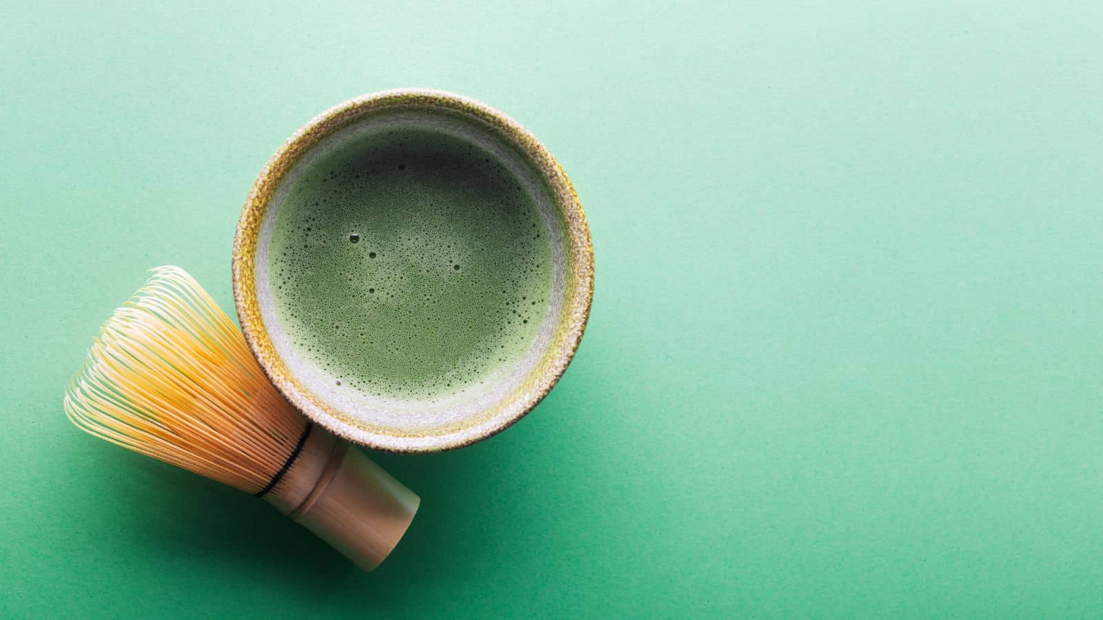 A ceramic bowl filled with frothy green matcha is placed on a green surface. Beside the bowl is a bamboo whisk, used for mixing the matcha. The image is captured from above, with the bowl positioned slightly to the left.