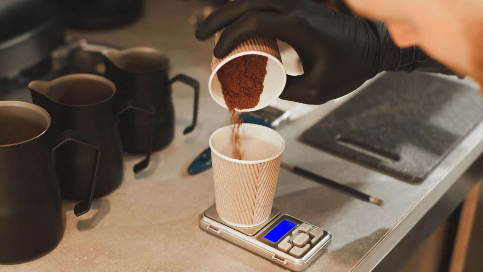 A person wearing black gloves pours ground coffee from a paper cup into another cup placed on a digital scale. Metal pitchers and a pencil are in the background on the countertop.