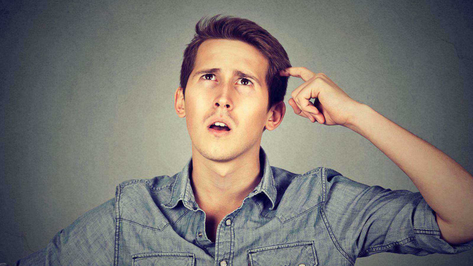 A young man with short brown hair is looking upwards with a thoughtful expression, touching his head with one finger. He is wearing a light blue denim shirt against a plain gray background.