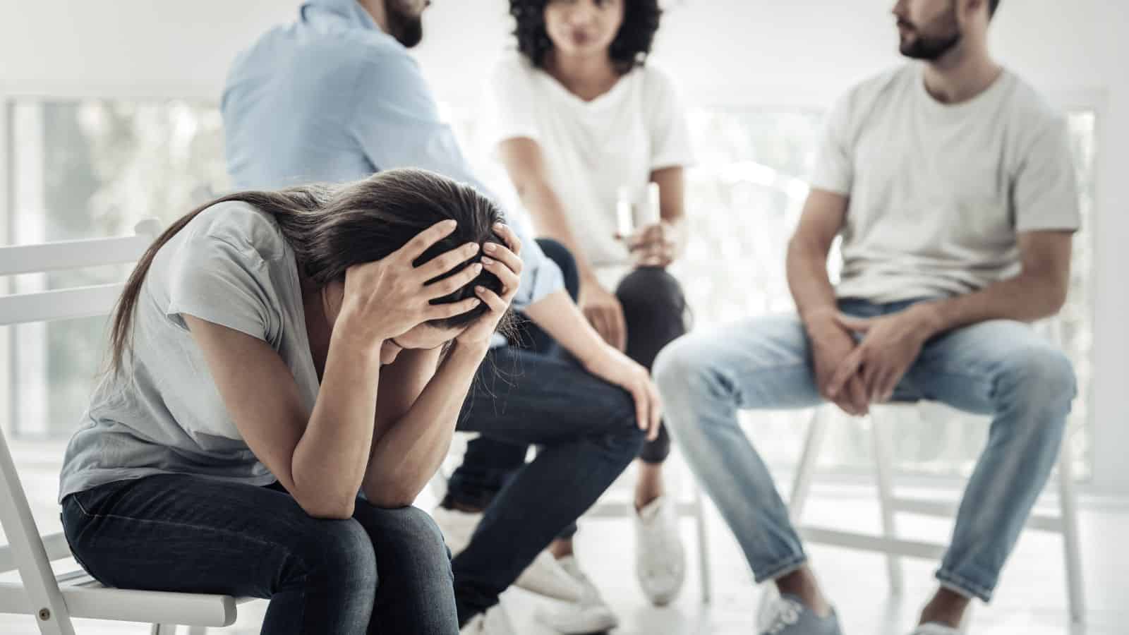 A woman sits holding her head in her hands, appearing upset, while three people sit nearby, engaged in conversation. The setting appears to be a group therapy session in a bright room with large windows.