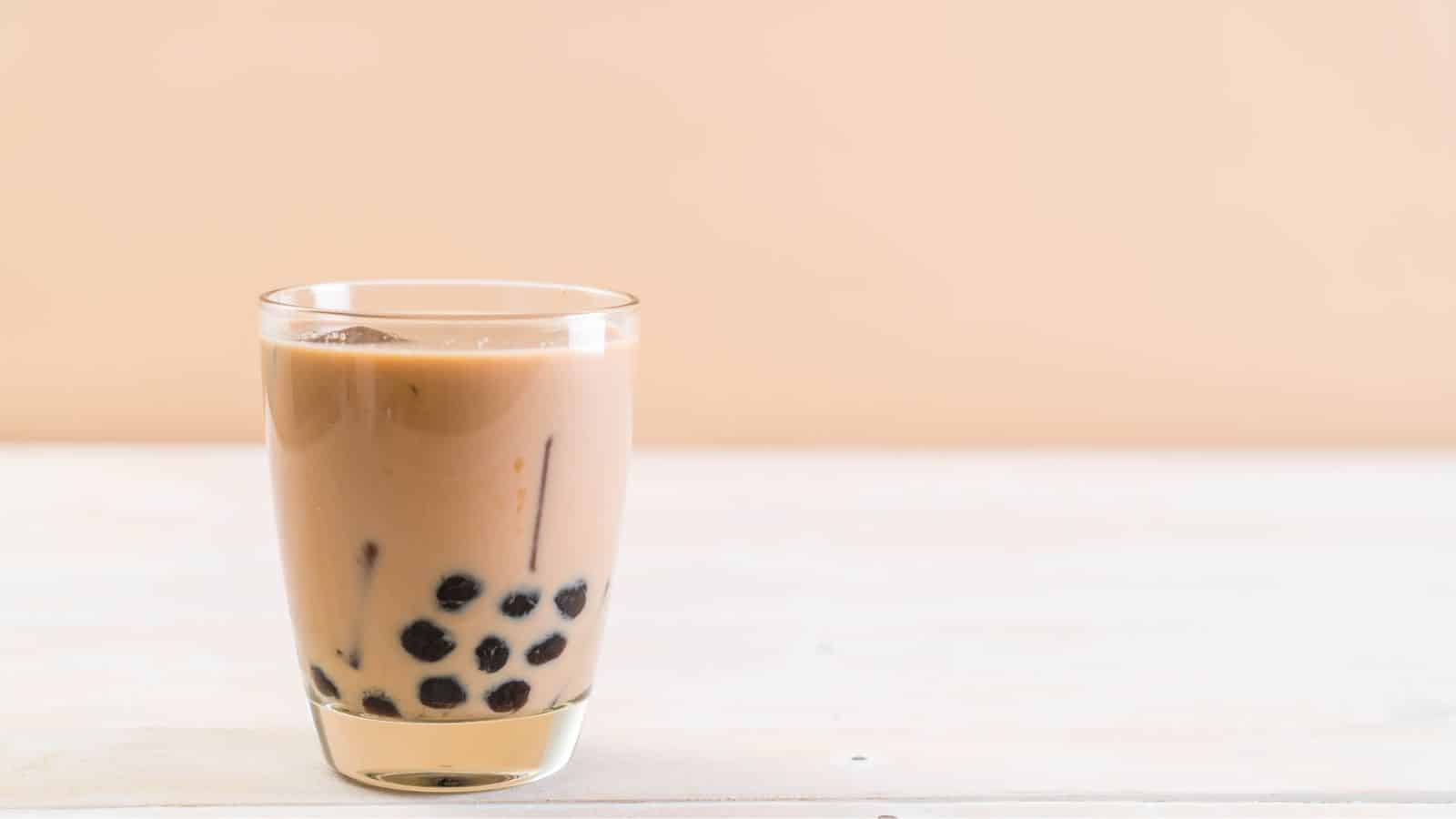 A glass of bubble tea with milk tea and black tapioca pearls sits on a light wooden surface. The background is a simple tan color.