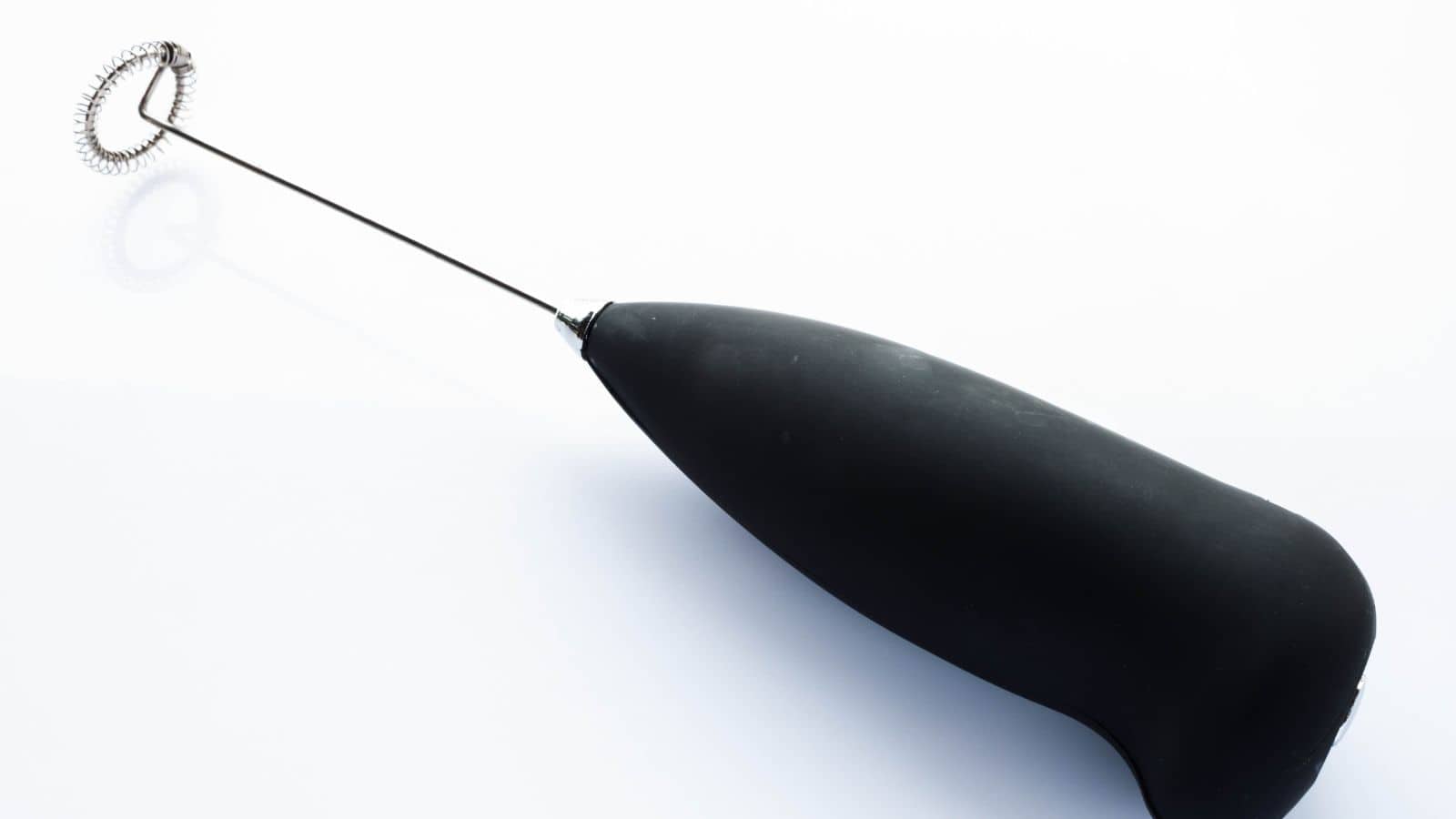A handheld milk frother with a black ergonomic handle and a thin metal whisk sits on a white surface.