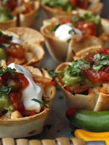 Mini taco cups on a baking sheet, filled with ingredients like guacamole, sour cream, salsa, and chopped herbs. Two chili peppers are in the foreground.