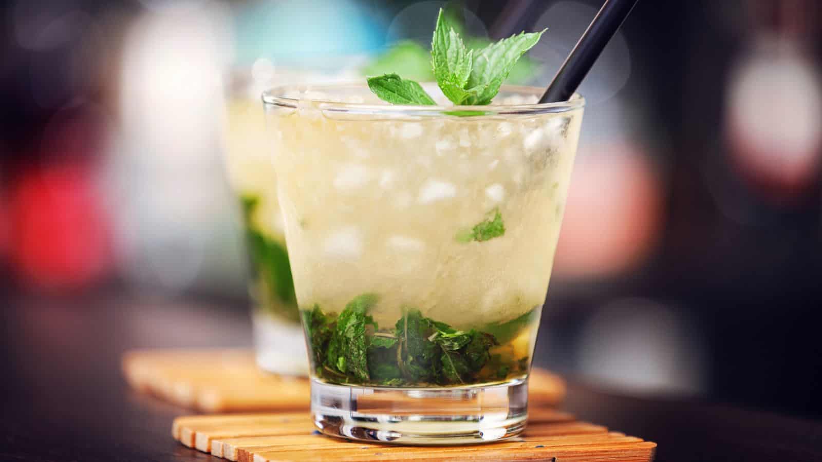 A glass of mint julep, featuring crushed ice, mint leaves, and a straw, sits on a wooden coaster. In the background, another identical glass is slightly out of focus. The setting appears to be a bar or similar establishment.