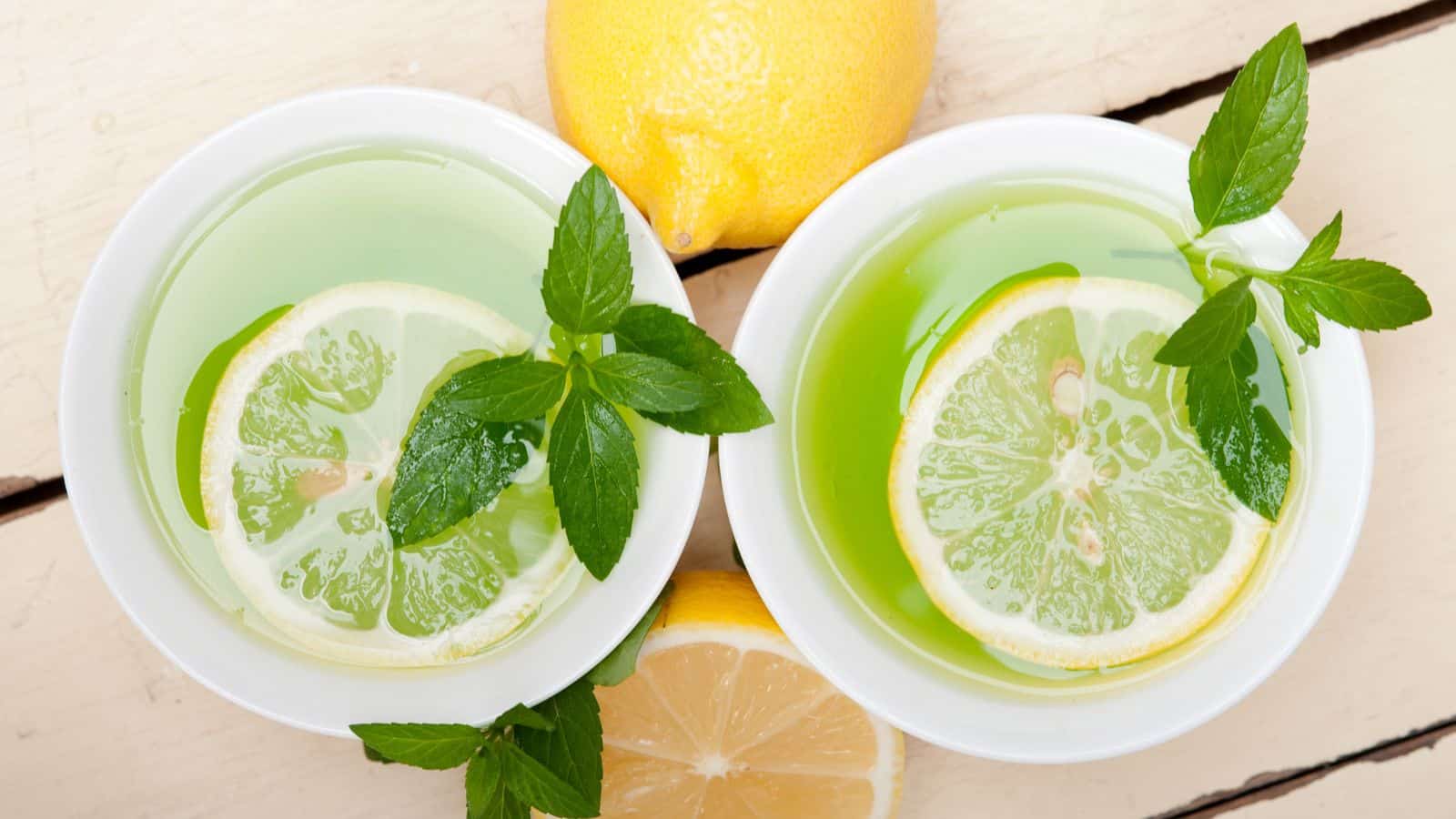 Top view of two white bowls filled with a green liquid, each garnished with lemon slices and mint leaves. A whole lemon and a half lemon are placed on a light wooden surface beside the bowls.