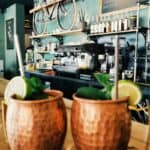 Two copper mugs filled with drinks, garnished with lime and mint, sit on a wooden counter. The background reveals a cafรฉ interior featuring a vintage bicycle mounted on the wall, a menu board, and various bottles on shelvesโessential elements for crafting must-have mocktail mixers.