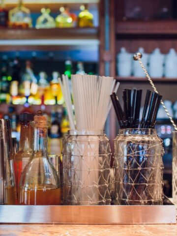 A must-have bar setup features metal shakers, a small bottle of bitters, containers with white and black straws, glass stirrers, and a cocktail glass with mint on a marble countertop. In the background, shelves are stocked with an array of liquor bottles and essential mocktail mixers.