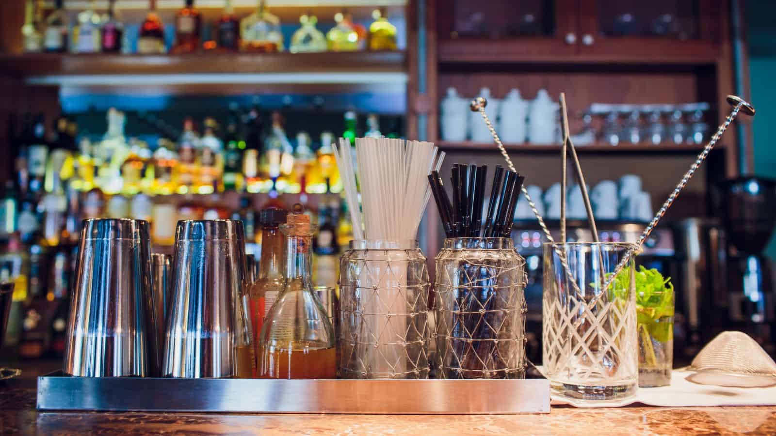 A must-have bar setup features metal shakers, a small bottle of bitters, containers with white and black straws, glass stirrers, and a cocktail glass with mint on a marble countertop. In the background, shelves are stocked with an array of liquor bottles and essential mocktail mixers.